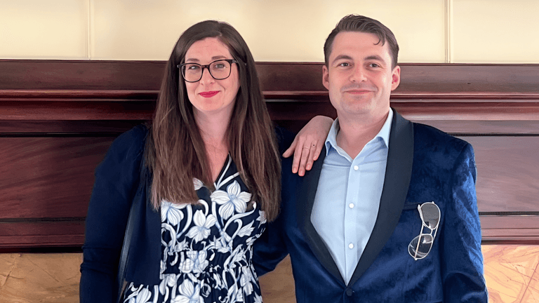 Man and woman smiling in front of wooden panel, dressed in business attire.