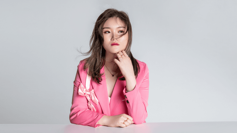 A woman dressed in a pink blazer with a bow, sitting at a table with a thoughtful expression. Her hair is slightly tousled, and she is gently resting her chin on her hand while looking to the side, against a simple gray background.