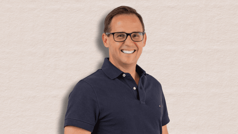 Smiling professional man in a navy polo shirt with glasses, standing against a soft beige background, exuding confidence and approachability.