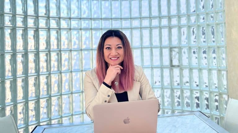 "Professional portrait of a woman with pink ombre hair, sitting at a desk with a MacBook, smiling and resting her chin on her hand, against a backdrop of a glass block wall."