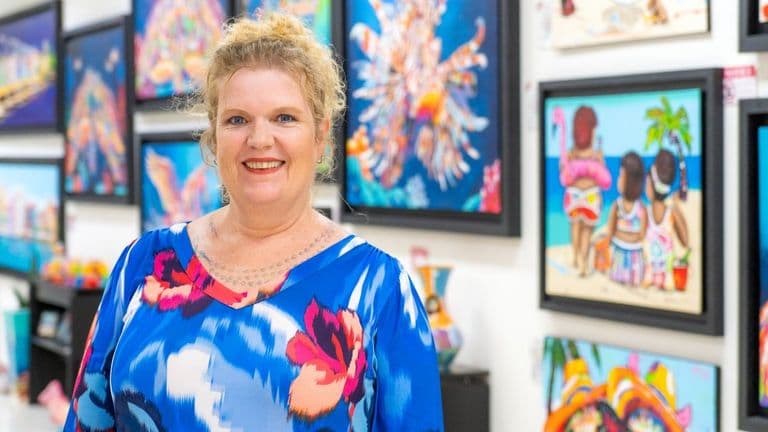 Portrait of a smiling artist standing in front of vibrant artwork featuring bright tropical designs and abstract art.