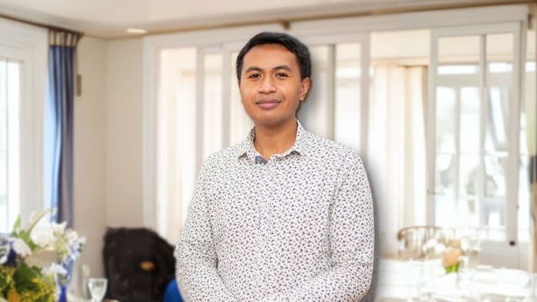 Man in a floral shirt standing indoors with a soft smile, surrounded by natural light and a blurred background of curtains and floral arrangements.
