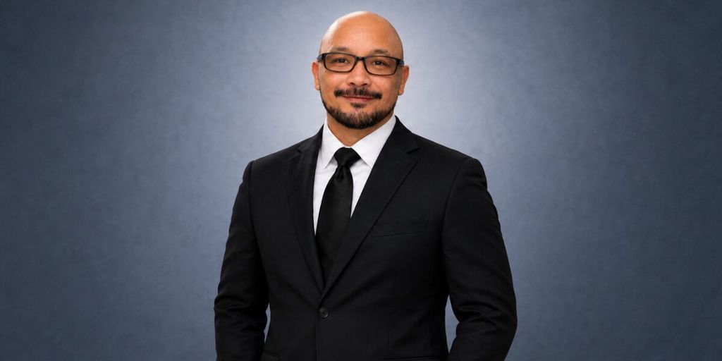 Professional portrait of Charles Santos wearing a black suit and tie against a gray background.