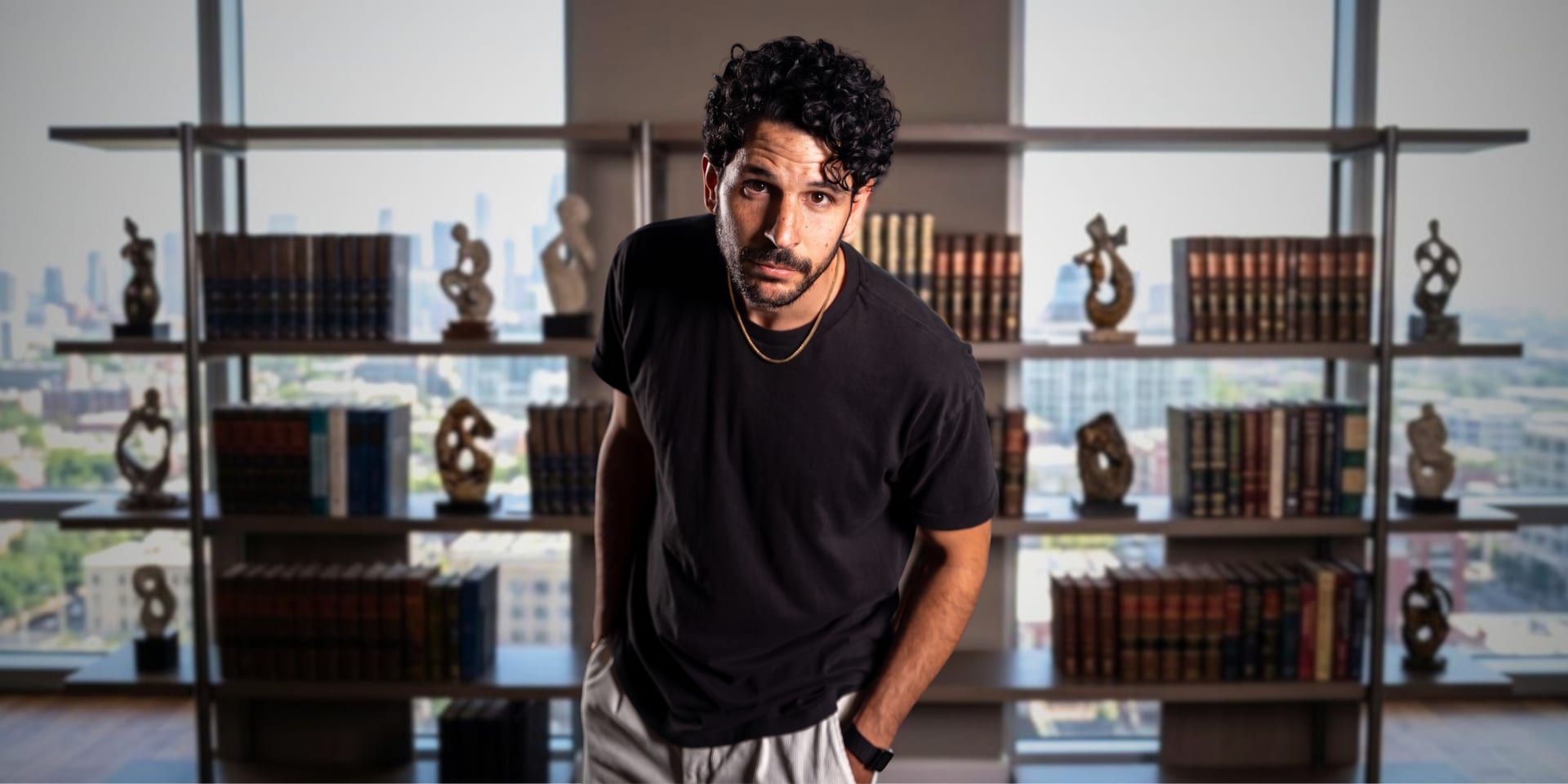 Man with curly hair, wearing a black t-shirt, standing in front of a bookshelf with a city view.