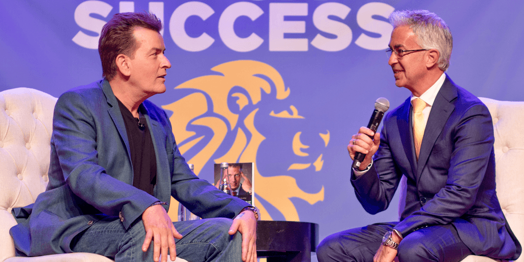 Craig Carr speaks on stage with another man during a business success event promoting psychology-based wealth and sales strategies.