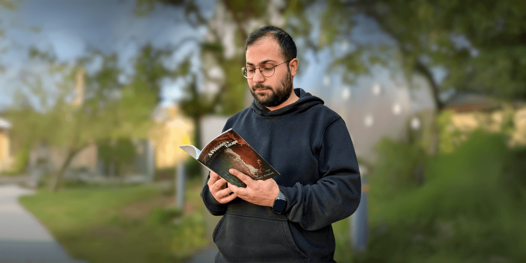 A man reading "A Prisoner's Escape" by Johnny Achkar outdoors, reflecting themes of resistance, exile, and self-reclamation.