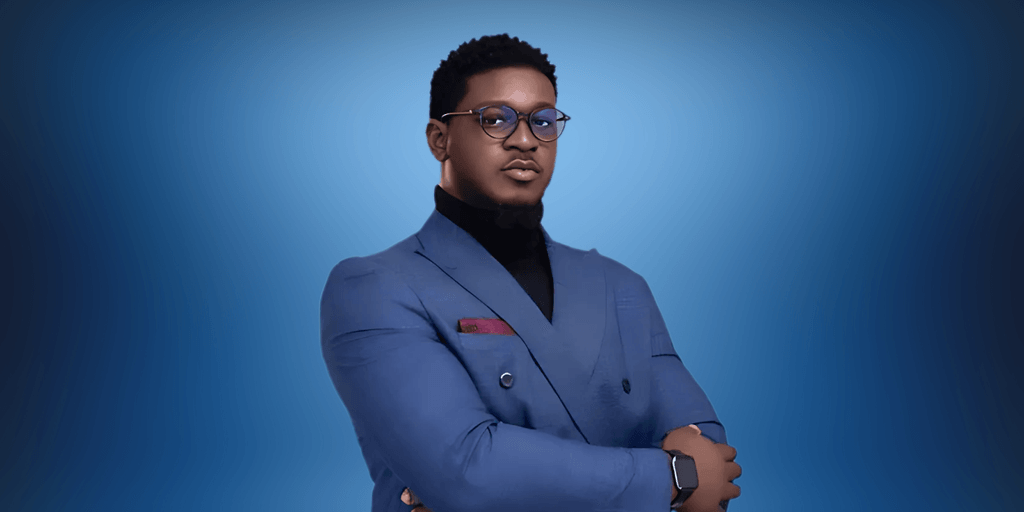 "Professional portrait of a man in a blue suit, wearing glasses, with his arms crossed confidently. The background features a gradient blue tone, creating a sleek and modern look."