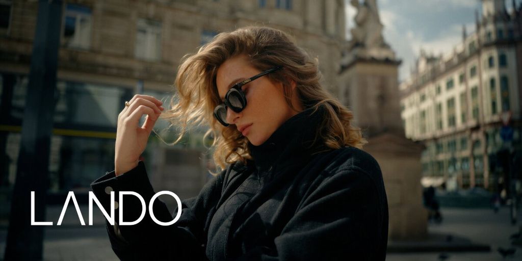A woman in sunglasses and a black coat stands in a city square, with elegant architecture behind her. She looks contemplative. The word "Lando" is displayed.