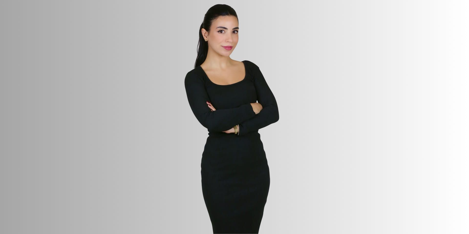 Woman in black dress standing confidently with arms crossed against a neutral background.