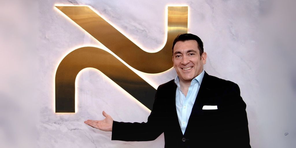 A man standing next to a glowing NovoSculpt logo on a marble wall, smiling and presenting the brand with a welcoming gesture. The logo represents the NovoSculpt fat removal technology.