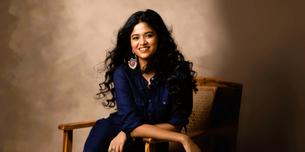 Smiling woman with curly hair in a navy blue dress and statement earrings, sitting on a chair in a relaxed pose, ideal for content related to lifestyle, beauty, and personal empowerment.