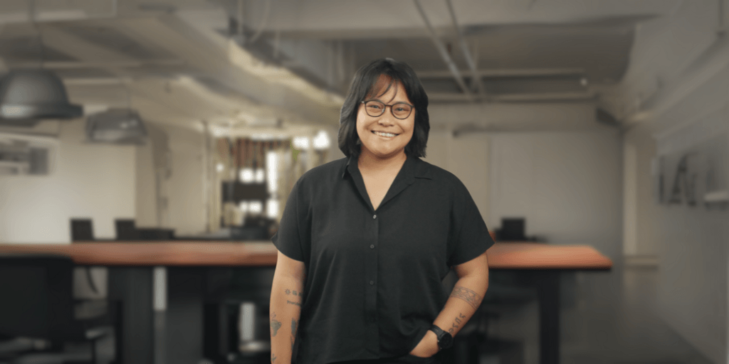 Portrait of a woman with short black hair and glasses, smiling confidently in a black button-up shirt, standing in an office setting with blurred background.