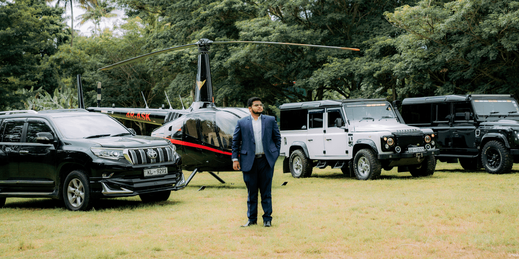 Man in a blue suit standing in front of a helicopter and a lineup of black and white off-road vehicles, set in a scenic outdoor location.