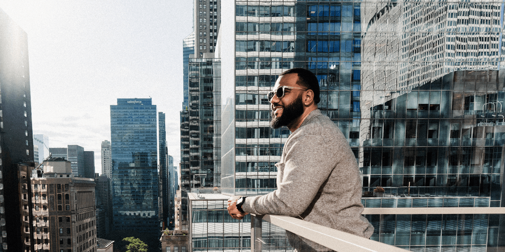 A man standing on a rooftop terrace with a cityscape background, smiling while wearing sunglasses, with modern glass buildings reflecting the sunlight.