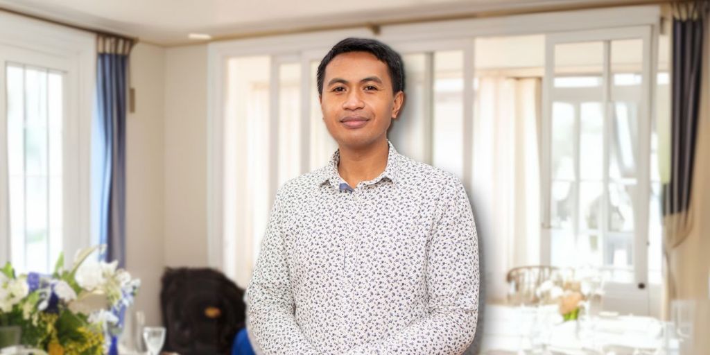 Man in a floral shirt standing indoors with a soft smile, surrounded by natural light and a blurred background of curtains and floral arrangements.
