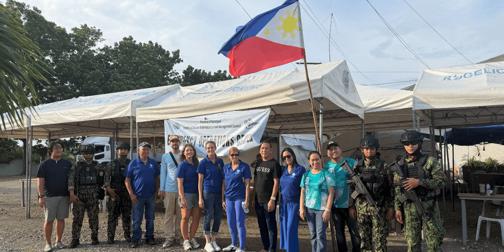Group of people in blue shirts with military personnel under Philippine flag and tents