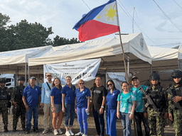 Group of people in blue shirts with military personnel under Philippine flag and tents