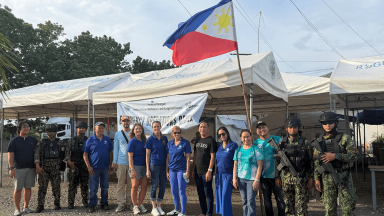 Group of people in blue shirts with military personnel under Philippine flag and tents