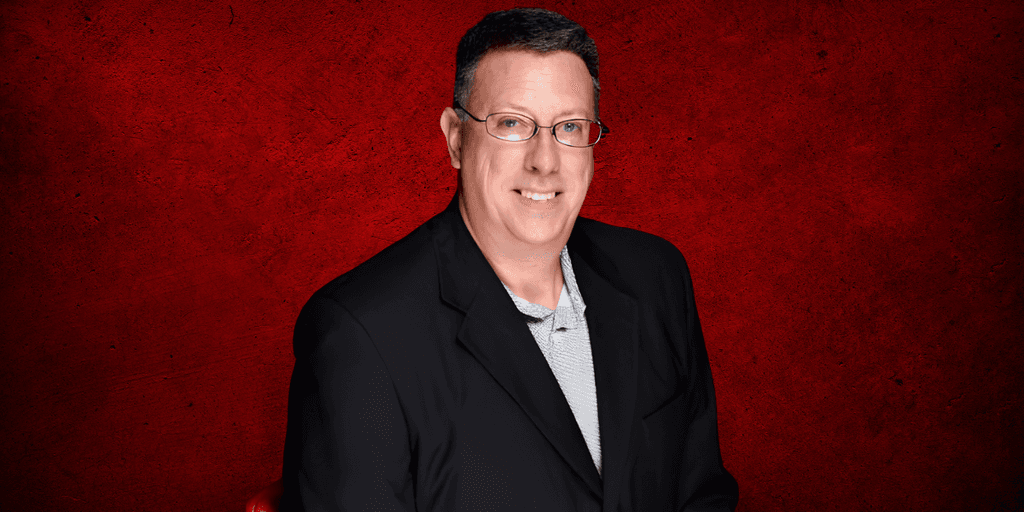 Man in a black blazer stands against a red textured background smiling