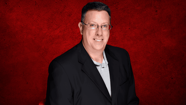 Man in a black blazer stands against a red textured background smiling