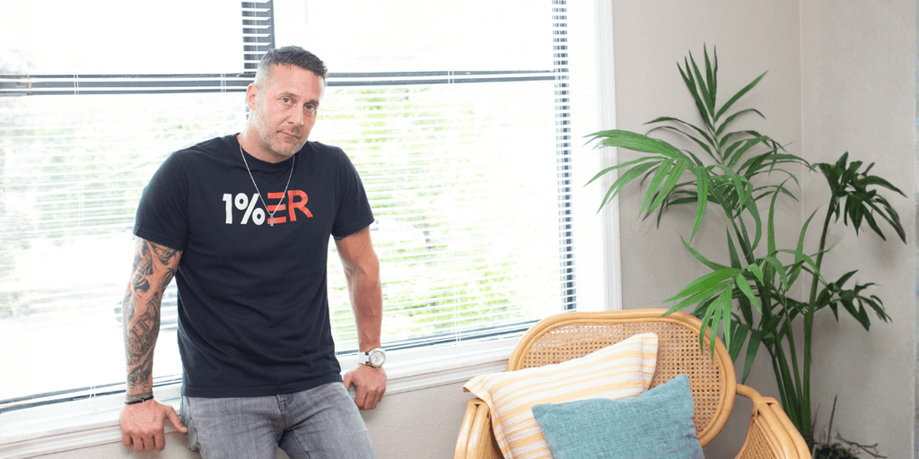 Man in a black t-shirt leans against a window with indoor plants and a chair