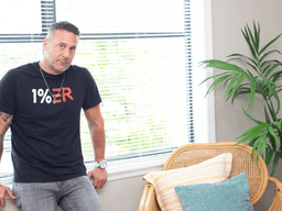 Man in a black t-shirt leans against a window with indoor plants and a chair