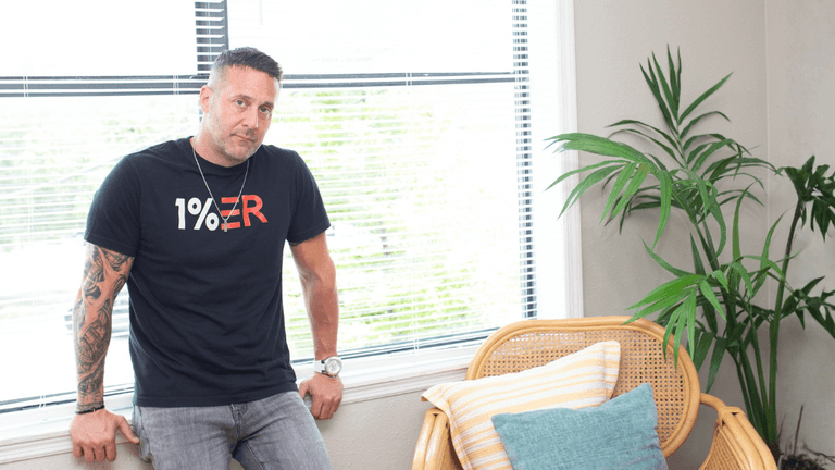 Man in a black t-shirt leans against a window with indoor plants and a chair