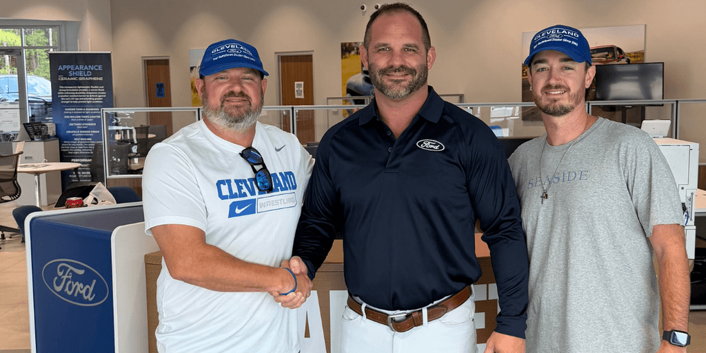 Three men shaking hands inside a Ford dealership, all smiling and wearing hats.