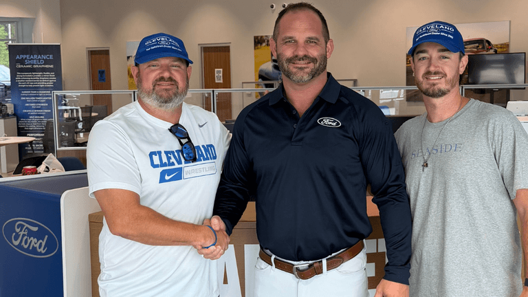 Three men shaking hands inside a Ford dealership, all smiling and wearing hats.