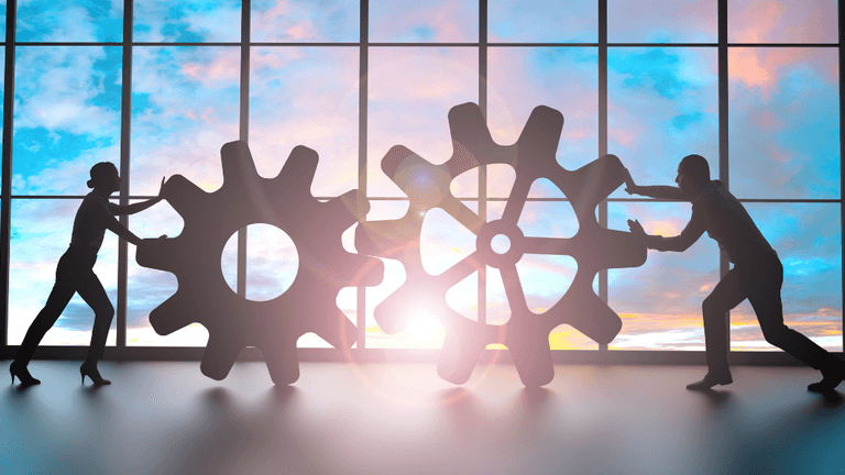 Silhouettes of a man and a woman pushing large gears against a sunset backdrop