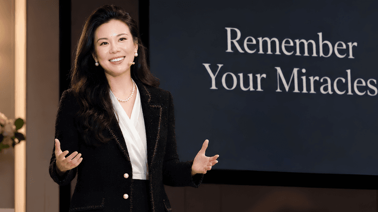 A woman in a black blazer speaks confidently in front of a presentation screen