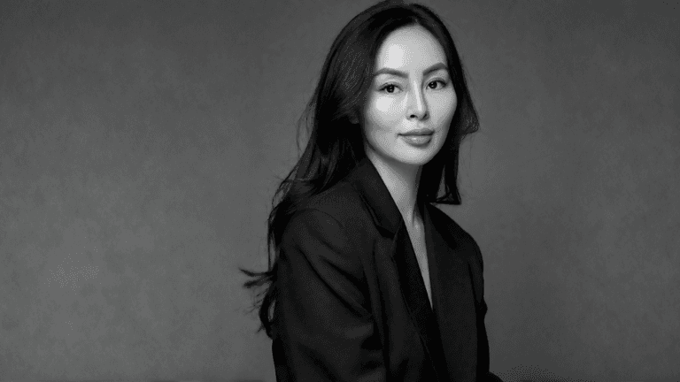 A woman in a black suit poses against a neutral gray background
