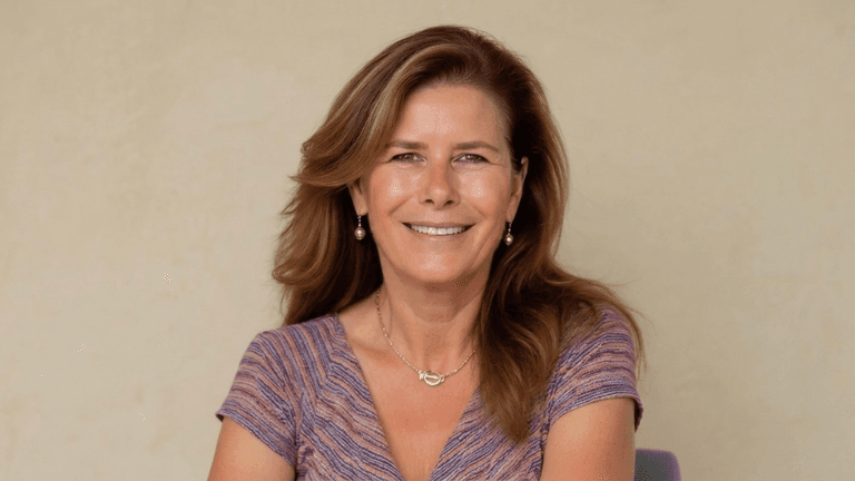 Woman with long brown hair smiles, wearing a colorful top and earrings