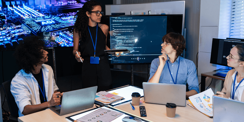 A diverse group collaborating in a tech meeting with digital presentations and coding on screens