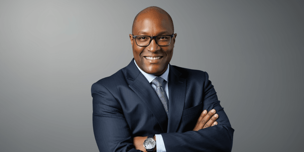 Smiling man in a suit with glasses, arms crossed, against a gray background