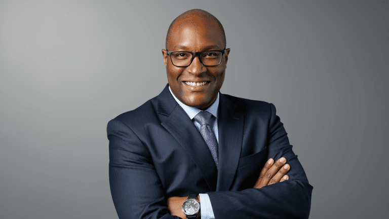 Smiling man in a suit with glasses, arms crossed, against a gray background