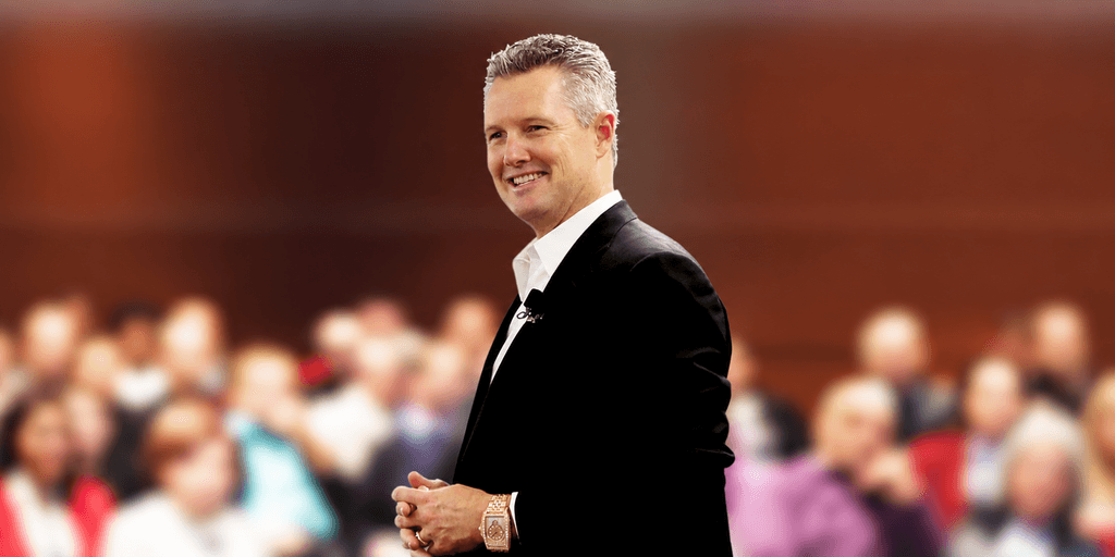A man in a suit smiles confidently in front of an audience.