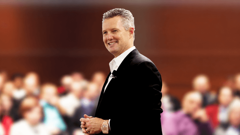 A man in a suit smiles confidently in front of an audience.