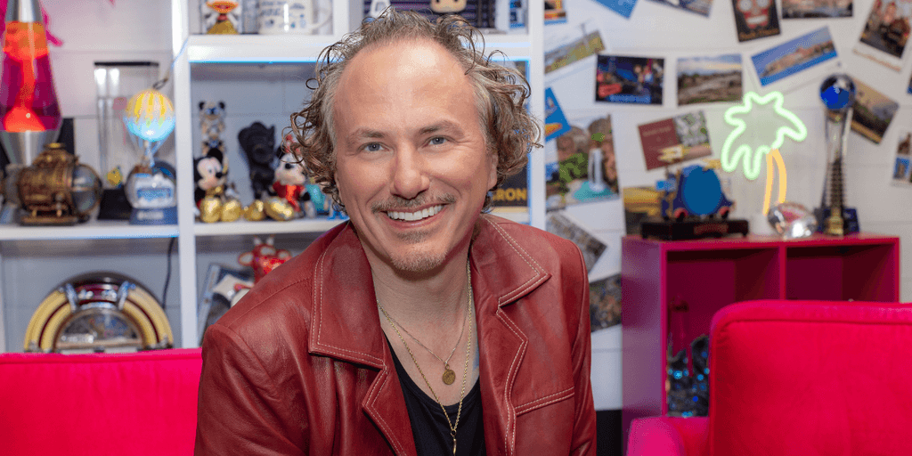 A smiling man in a red jacket poses against a colorful, playful background