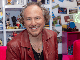 A smiling man in a red jacket poses against a colorful, playful background