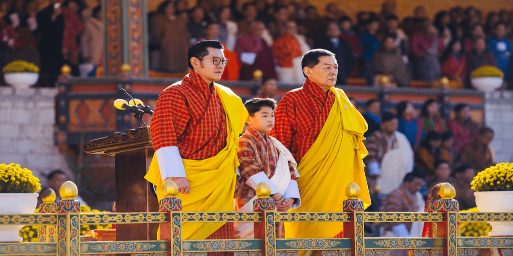 Three Bhutanese individuals in traditional attire stand at a ceremonial podium.