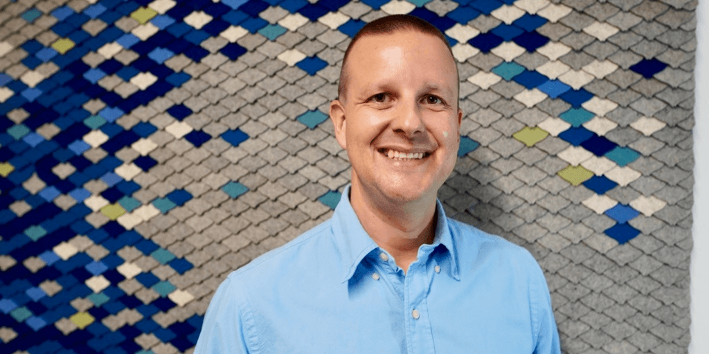 Smiling man in a blue shirt stands in front of a textured blue wall.