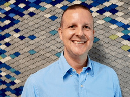 Smiling man in a blue shirt stands in front of a textured blue wall.