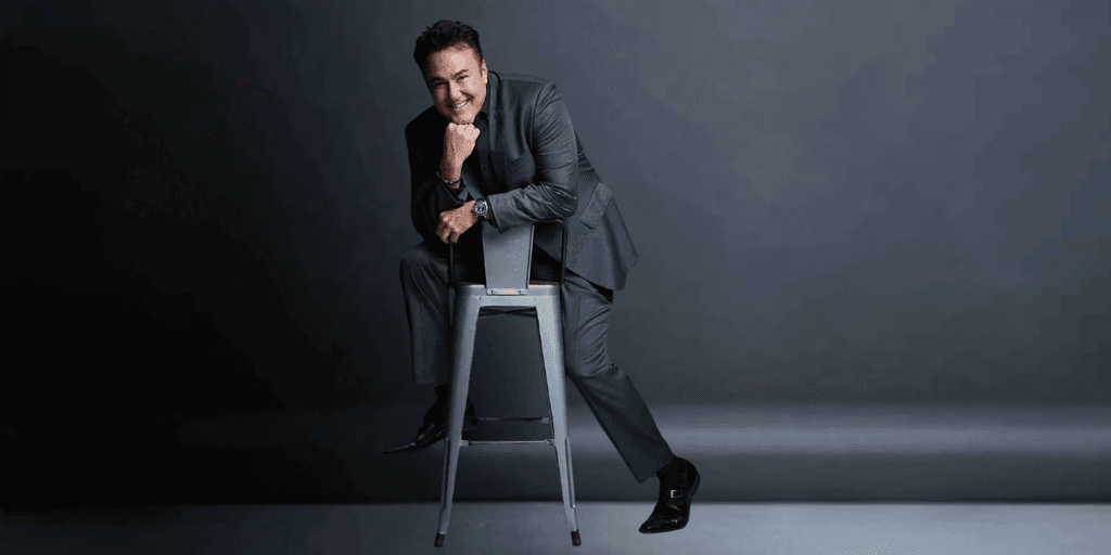 A smiling man in a suit poses playfully on a stool against a dark background