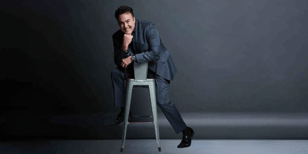 A smiling man in a suit poses playfully on a stool against a dark background