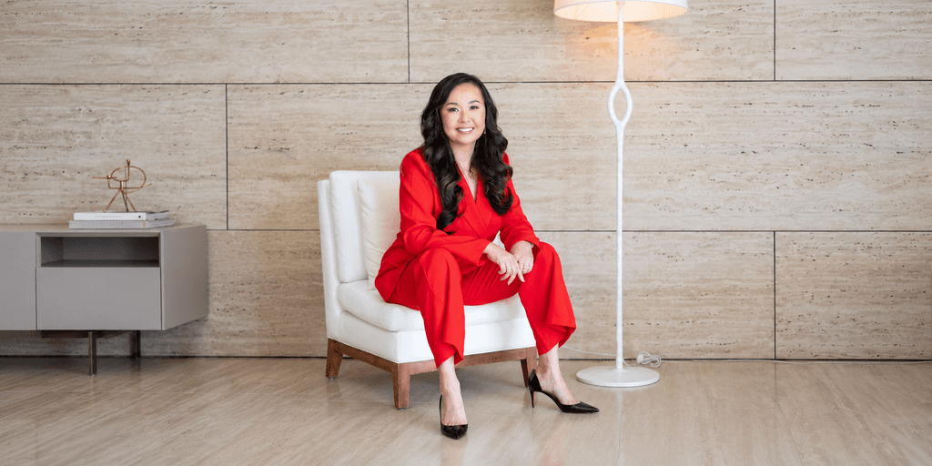 A woman in a red suit sits confidently on a white chair indoors