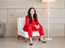A woman in a red suit sits confidently on a white chair indoors