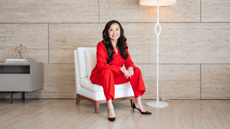A woman in a red suit sits confidently on a white chair indoors