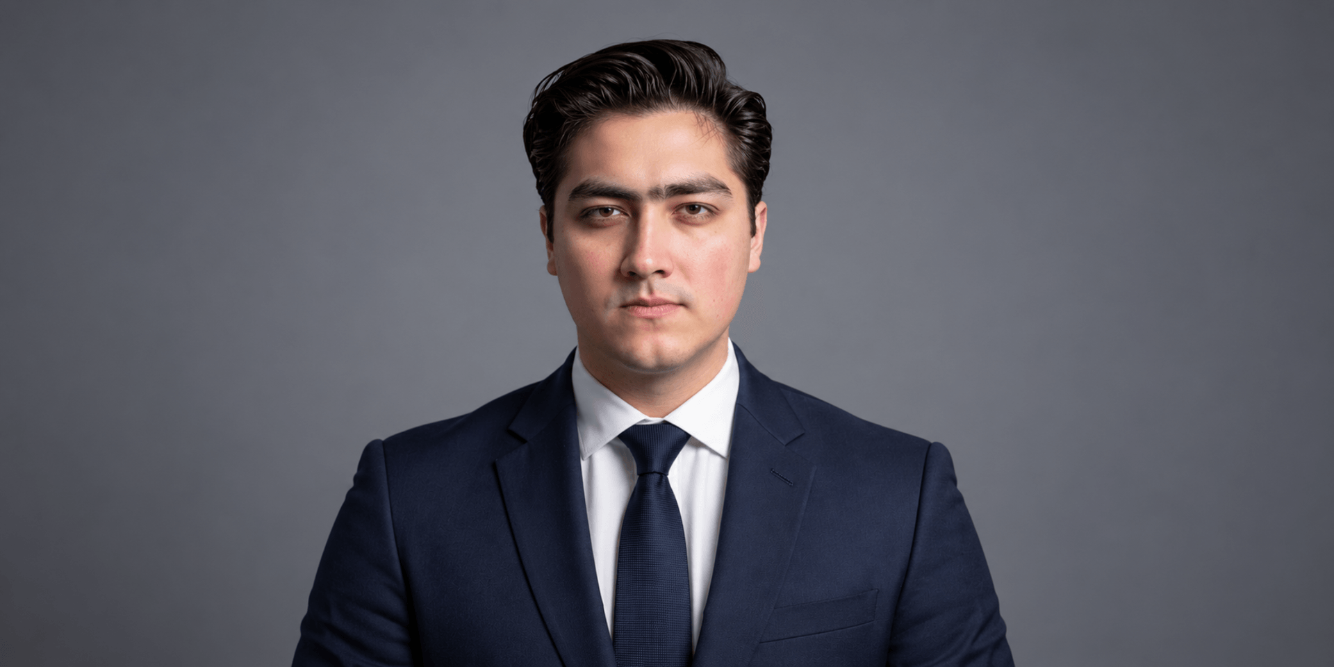 A serious young man in a navy suit and tie against a gray background