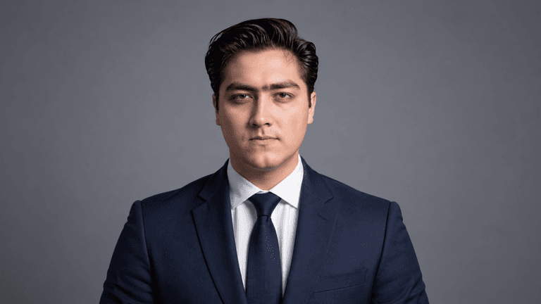A serious young man in a navy suit and tie against a gray background